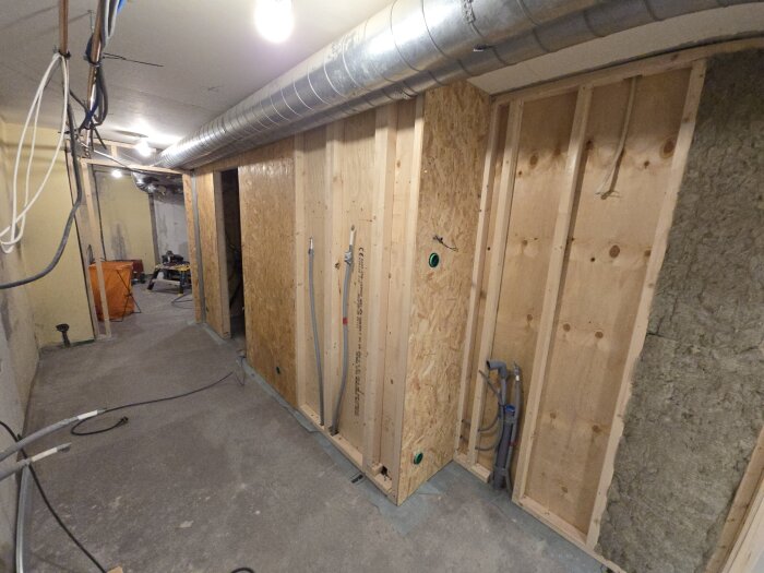 En pågående källarrenovering med träreglar, OSB-skivor och synliga rör, fläktrör i taket och oisolerade väggar, samt belysning i taket.
