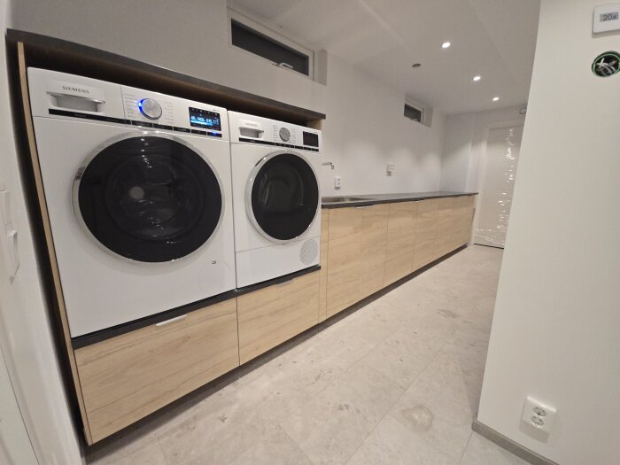 Tvättstuga med tvättmaskin och torktumlare, samt långa träskåp och diskbänk i modernt renoverad källare.