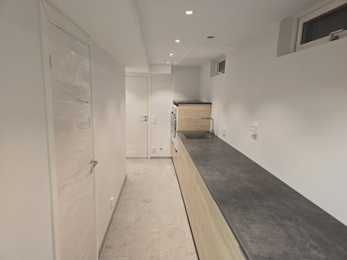 Nyrenoverad källare med köksbänk, diskho och ljusa väggar. Dubbeldörrar täckta med plast i bakgrunden.