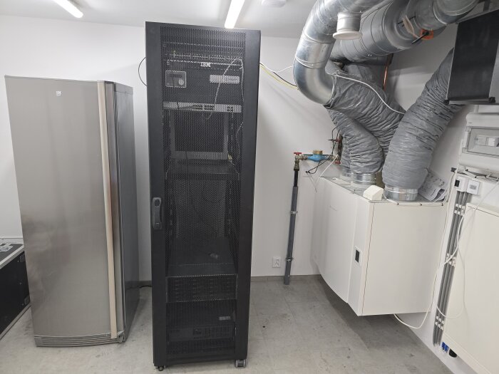 Serverrack, kylskåp och ventilationssystem i ett tekniskt rum med LED-takbelysning, synliga rör och kablar på väggarna.