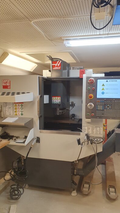 CNC-maskin i ett garage, märkt Haas, med öppet arbetsområde och kontrollpanel, omgiven av rör och verktyg i en upplyst miljö.