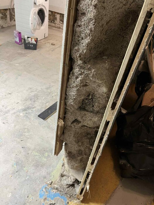Vägg med ojämn yta och håligheter i en källare under renovering, med exponerade reglar och en tvättmaskin i bakgrunden.