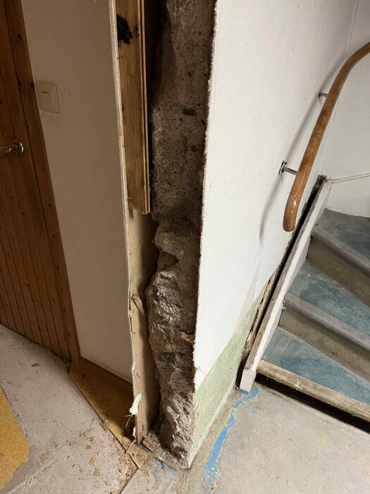 Ojämn bärande vägg i källaren från hus byggt 1934, delvis lös med synliga håligheter. Trävägg bredvid och trappräcke intill.