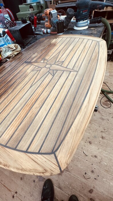 Mellanslipat teakbord med dekorativ design på ovansidan på en arbetsbänk omgiven av verktyg som slipmaskiner och skruvmejslar.