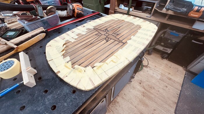 Teakbord med trästjärnmönster, maskeringstejp runt kanterna; verktyg och material på arbetsbänk, redo för vidare bearbetning.