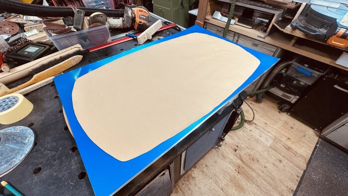 Mall av papper på en aluminiumplåt på ett arbetsbord, omgiven av verktyg som pensel, maskeringstejp och sandpapper.