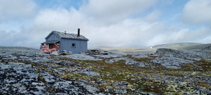 En liten stuga med kort skorsten på en karg bergssluttning i fjällen, under en molnig himmel.