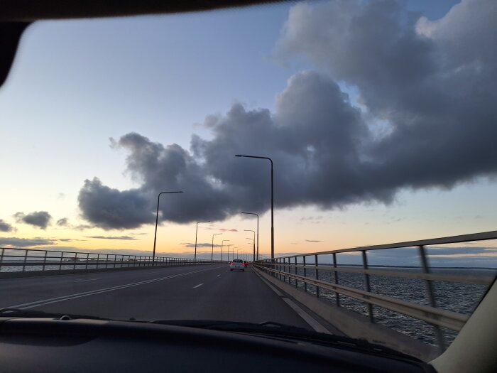 Bilfärd över lång bro med hav och solnedgång i bakgrunden, moln över himlen och flera gatlyktor längs vägen.