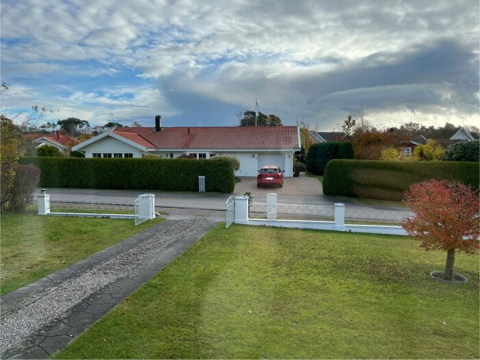 En villa med rött tak och garageuppfart, omgiven av häckar och ett lågt vitt staket, sedd från en gräsmatta och grusgång.