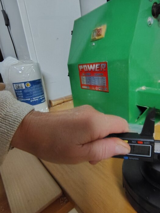 En hand håller ett digitalt skjutmått nära en grön maskin med märket "POWER" på en röd etikett.