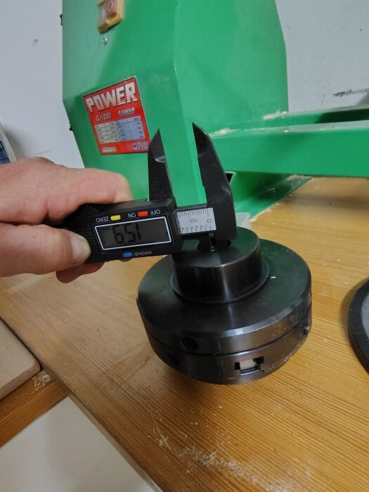 Digital skjutmått som mäter dimensionerna på en chuck i en verkstadsmiljö. En grön maskin är i bakgrunden.