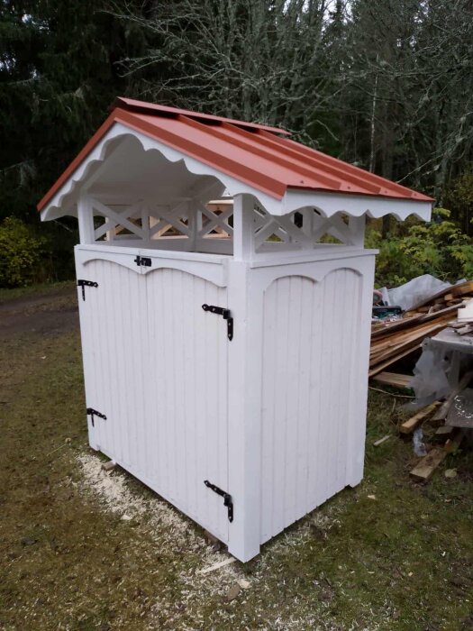 Vit soptunneskydd med rött tak, placerad på en gräsyta, omgiven av träd och träplankor i bakgrunden.