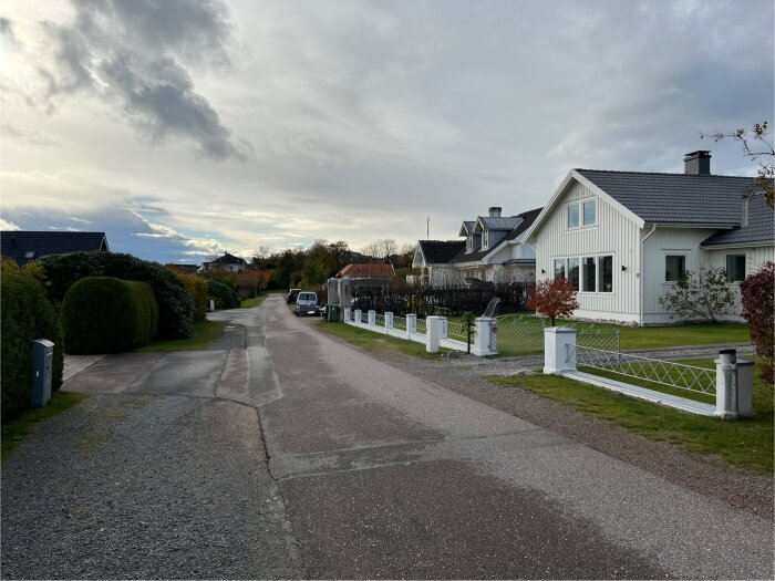 En lugn villagata med vita hus och trädgårdar till höger. Asfalterad väg med gräskant och buskar längs vänster sida av gatan.