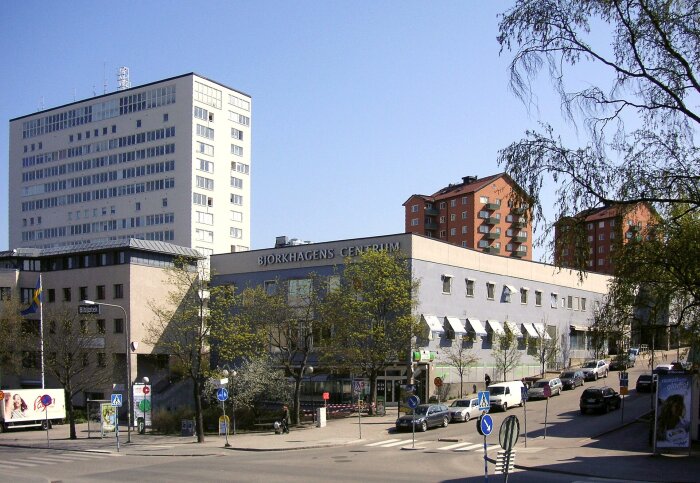 Gatubild av Björkhagens centrum med ett högre lägenhetshus i bakgrunden och flera parkerade bilar längs gatan.