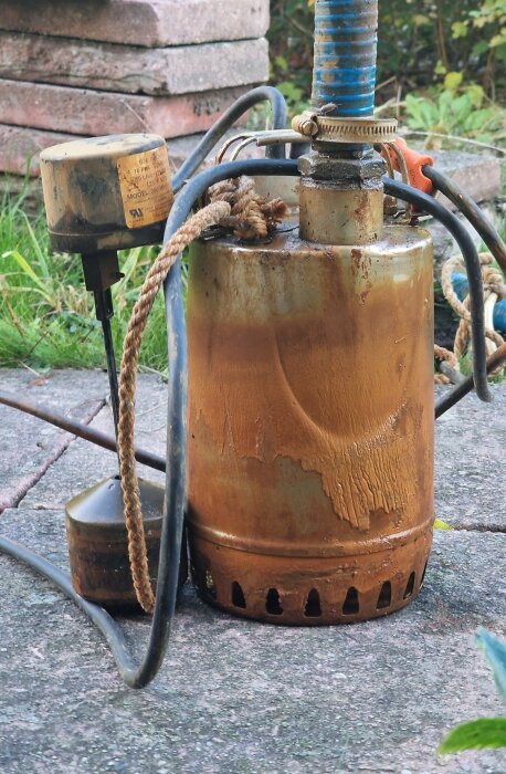 Rostig dräneringspump med elkablar och slang, placerad på stenplattor. En etikett med modellinformation är synlig på pumpen.