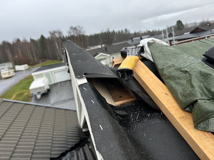 Tak med presenning och verktyg under byggnation, vy över bostadsområde i bakgrunden.