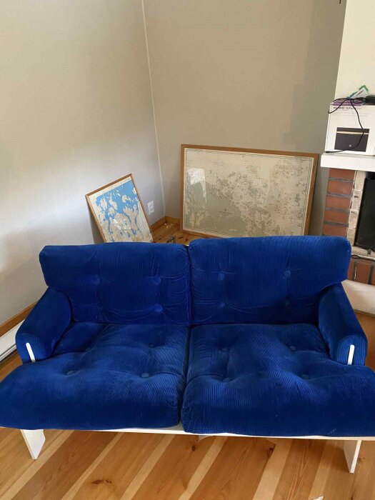 Blå sammetssoffa från 70-talet, stående i ett rum med trägolv, bredvid två kartor inramade på golvet och en öppen spis.