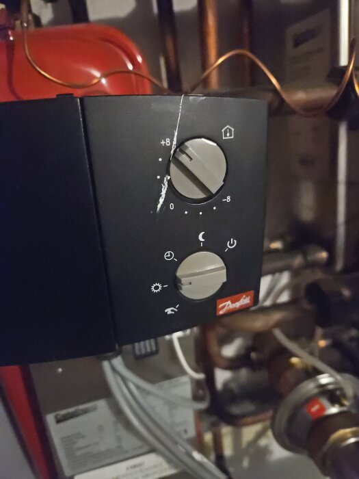 Termostatpanel från Danfoss med två vred, ett med temperatur och ett med symboler för dag och natt; rör och värmeutrustning i bakgrunden.