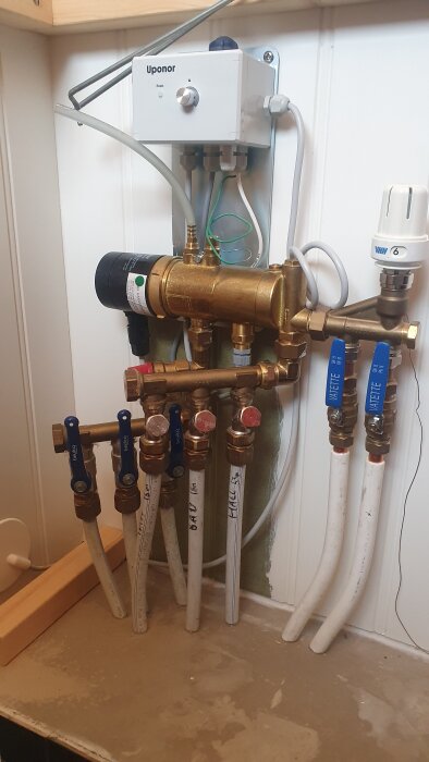 Cirkulationspump och ventiler för golvvärmeinstallation med rör och styrbox monterade på en vägg.