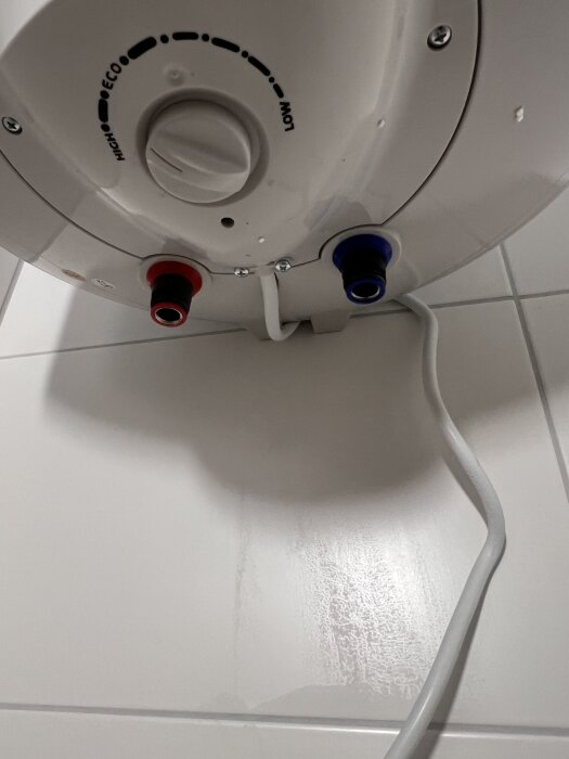 Varmvattenberedare med inställningsvred för temperaturnivåer, och röranslutningar för varmvatten (röd) och kallvatten (blå) mot kaklad vägg.