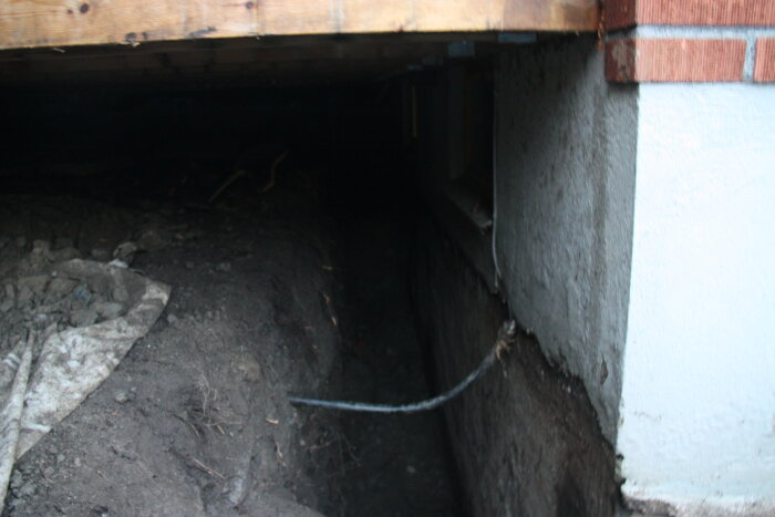 Under ett uterum, ett smalt och djupt dike grävt intill en vägg för dränering. Bakom, skymtas delar av byggkonstruktionen.