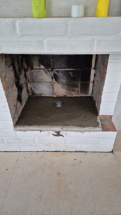 Nybetongad plattform i en öppen spis, omgiven av tegel och vitputsad mur, med ett rör infällt i betongen.