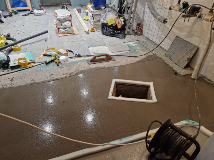 Flytspacklat källargolv med synlig inspektionslucka och omkringliggande byggmaterial. Verktyg och material sprids på sidan.