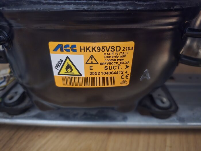 Närbild på en kompressor med ett gult klistermärke innehållande varningar och teknisk information för refrigerant R600a, tillverkad i Italien.