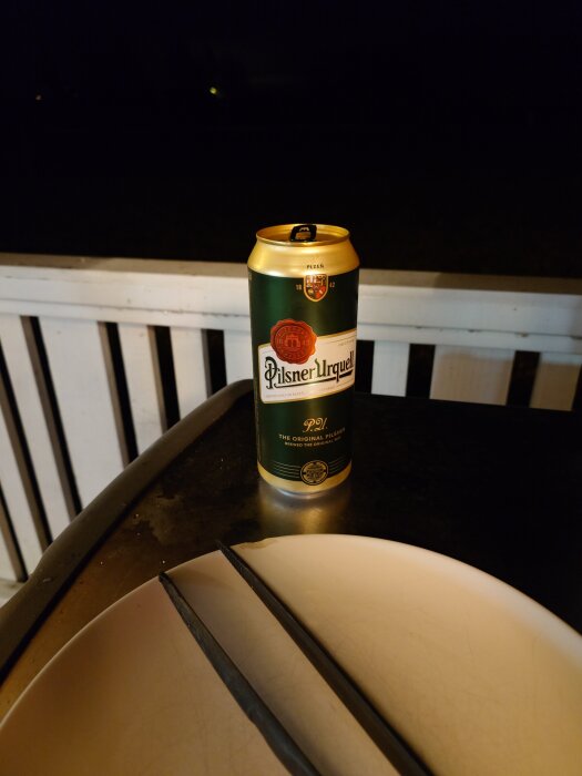 En burk Pilsner Urquell öl står på en grill med en tallrik och grilltång, kvällsmörker i bakgrunden.