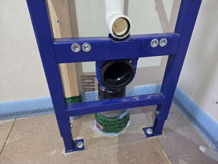 Blå ram för vägghängd toalettinstallation syns med röranslutning och träkonstruktion som stöd vid montering. Rörgenomföring vid golvet.