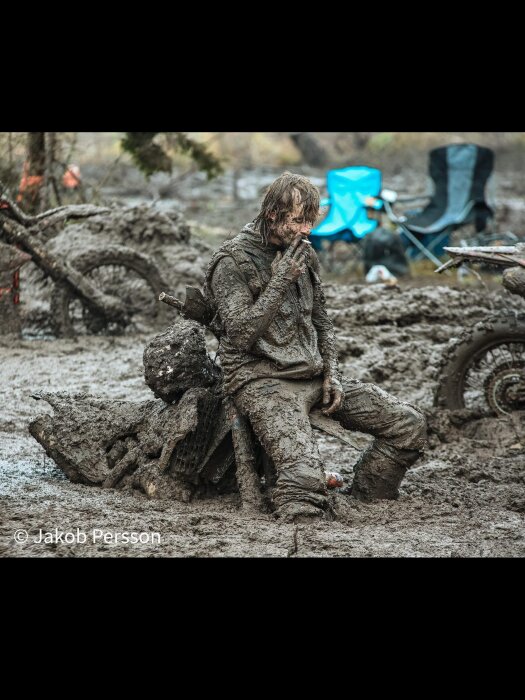En person täckt av lera sitter på en motorcykel omgiven av ett lerigt landskap med stolar i bakgrunden.