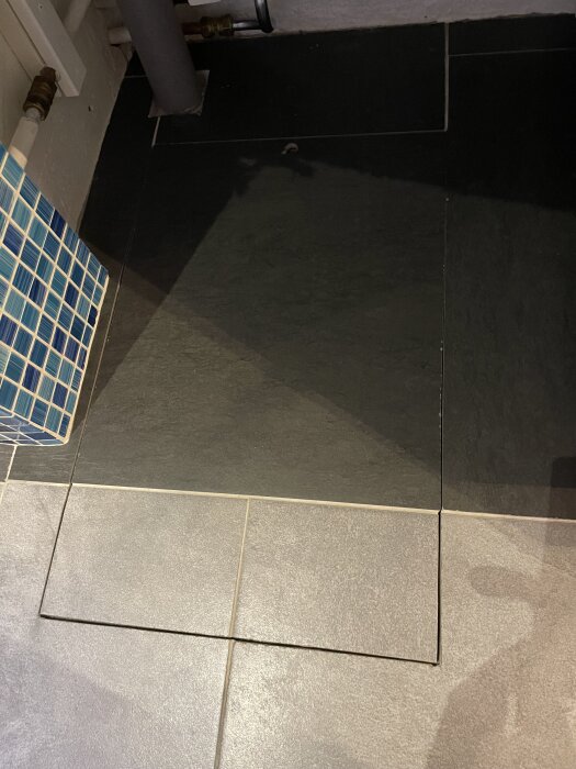 Golvyta med en lucka täckt av plattor, intill ett rörsystem och vägg med blå mosaikplattor.