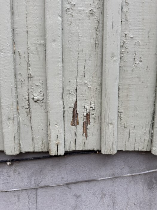 Fasadbrädor med skadad och flagnande färg samt små sprickor i träet vid husgrund.