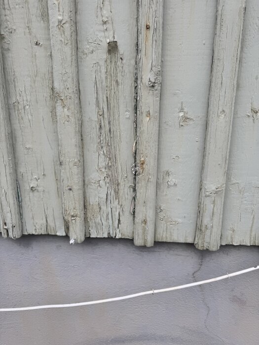 Fasadplankor med små sprickor i färgen, antydan till flagnad färg och synliga träytor. Plankorna sitter ovanför en gråmålad yta med ett vitt snöre.