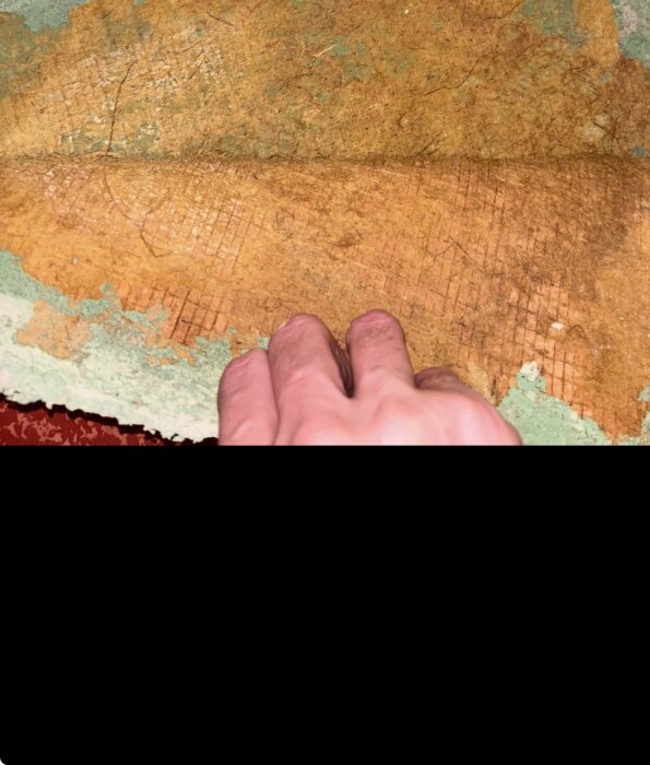 Hand drar upp gammal plastmatta i källare, avslöjar orange och grön yta under, misstänkt asbest från 1970-talet.