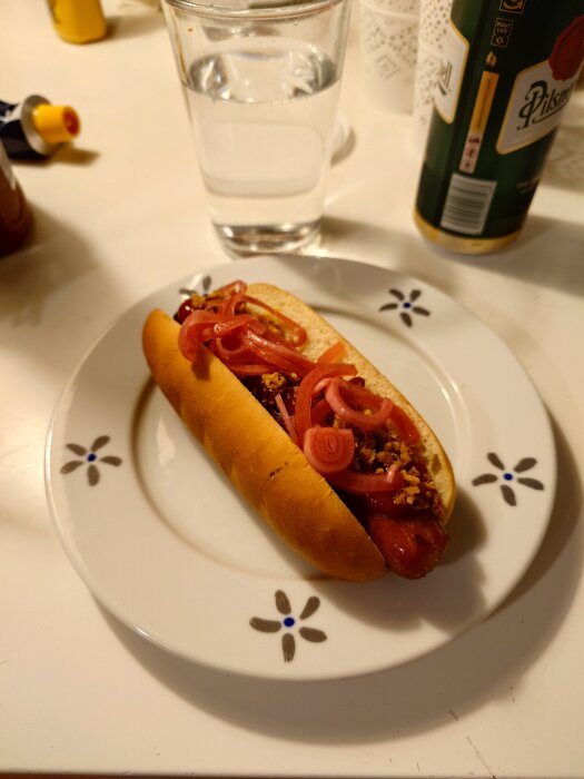 En grillad korv med bröd toppad med rostad och picklad lök på en vit tallrik, bredvid ett glas vatten och en burk öl.