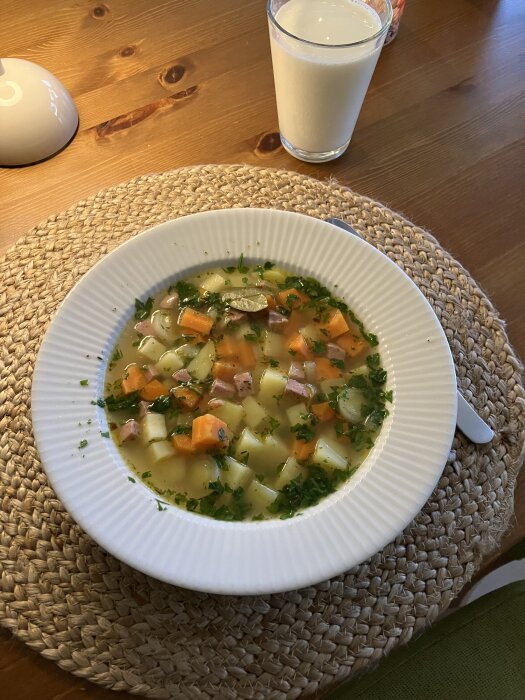 Grönsakssoppa med potatis och morötter i en vit tallrik på ett brunt bord, serverad med ett glas mjölk bredvid.