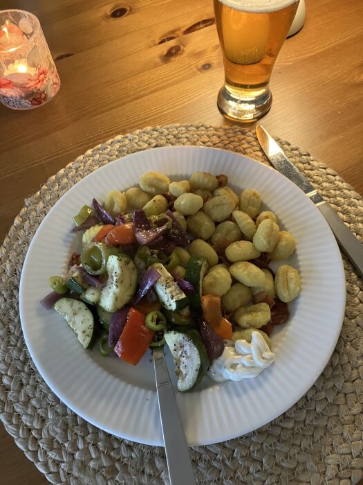 Tallrik med gnocchi, bacon, ugnsrostad zucchini, röd paprika, rödlök och sivripaprika, med ett glas öl vid sidan.