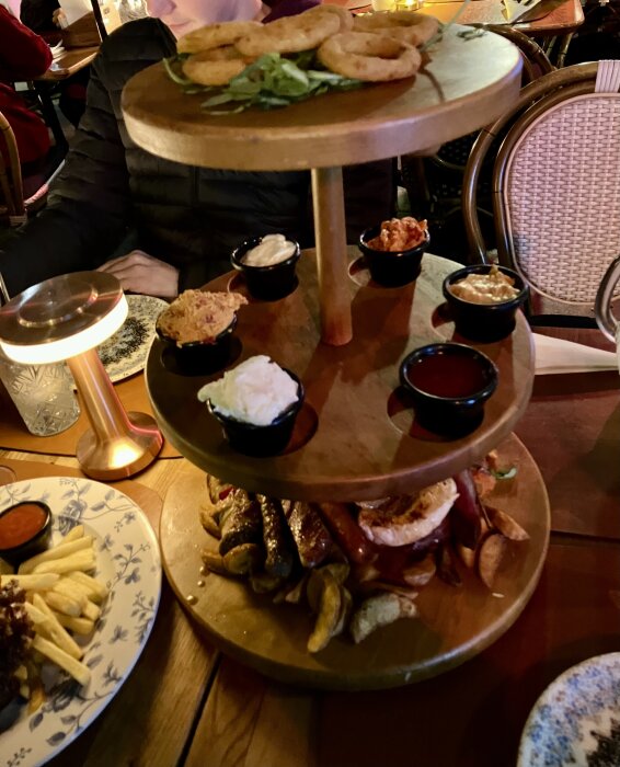 Tre våningsfat med olika snacks som lökringar, dippar och korvar på restaurangbord.