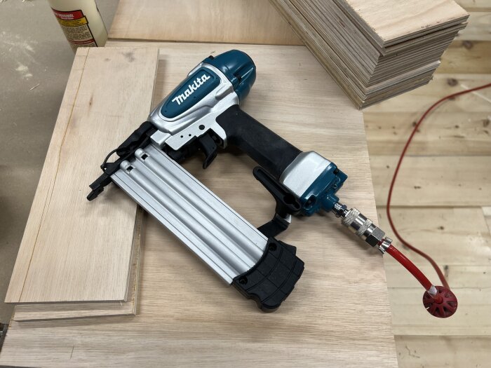 En ny Makita dyckertpistol pÄ en trÀyta, med staplade trÀplankor och en ansluten röd luftslang.