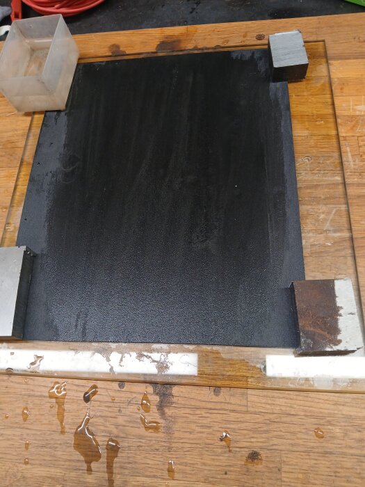 Parallellbitar av kalldragen plattstång på en glasskiva med våtslippapper, omgivna av träyta och vattenstänk.