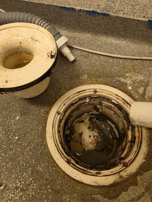 Äldre golvbrunn i tvättstuga med smuts och rost runt kanterna, delvis isig och ovanligt vattenrör synligt på en betonggolvyta.