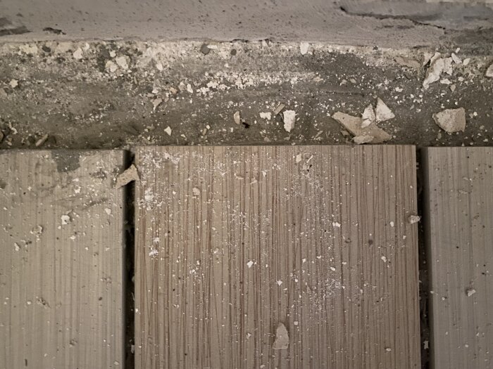 Klinkerplattor placerade löst på ett golv, med fogar synliga och byggdamm ovanpå. Kanten mot väggen är också fylld med byggdamm.