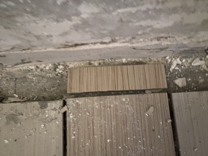 Närbild på golv där klinkerplattor ligger löst mot väggen, med damm och murbrukspartiklar på ytan.