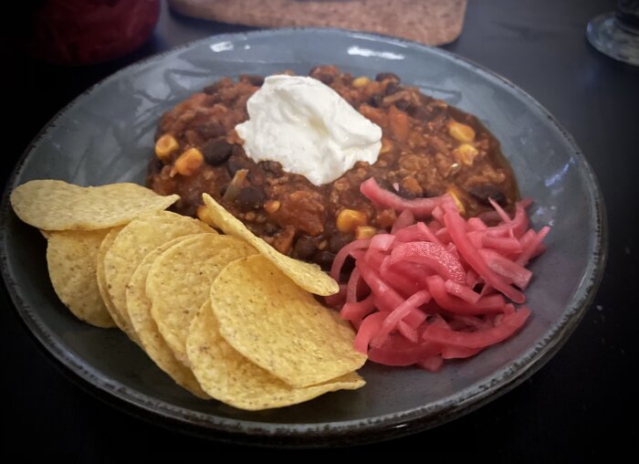 Chili con carne med gräddfil, serverad med nachos och picklad rödlök på en tallrik.