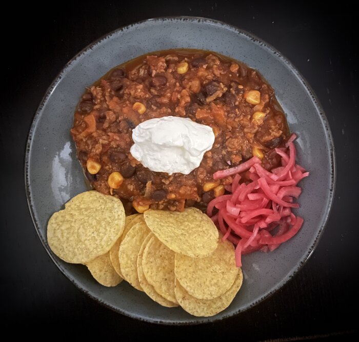 Chili con carne med gräddfil, nachos och picklad rödlök serverat på en tallrik.
