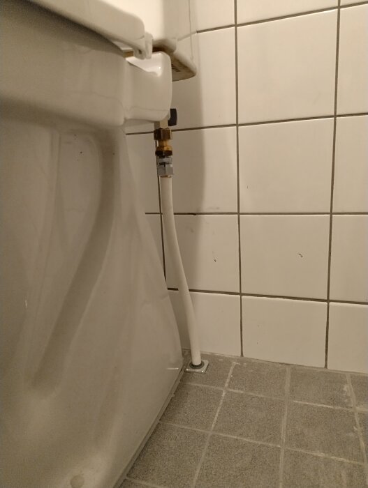 Kopparrör med plastisolering leder till en toalett bredvid vita kakelplattor på en grå våtrumsmatta.