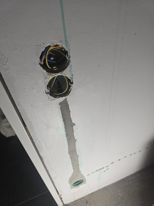 Vägg med två eluttag i olika hål, synliga kablar, spår av cement och en vertikal linje av cementputs och gröna markeringar.