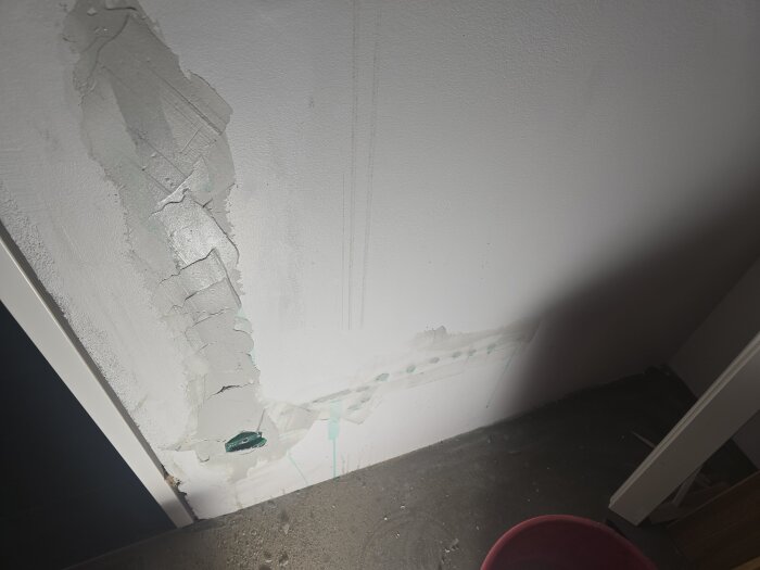 Vägg med spacklade reparationer och ett grönt eluttag som syns vid nedre vänstra hörnet. Golvet har fragment av målararbete.