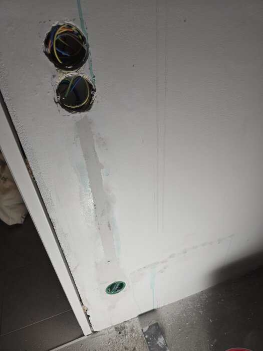 Vägg med två öppningar för eluttag, synliga kablar, gröna markeringar och nyligen applicerat spackel, redo för grundmålning.